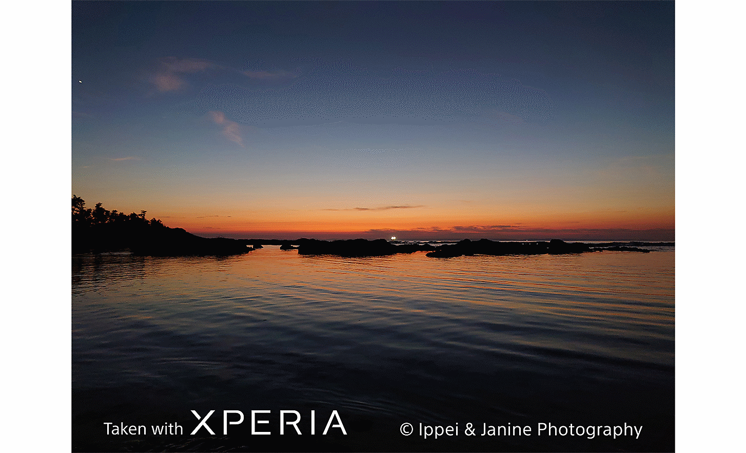 Slnko zapadajúce za veľkú vodnú plochu. Na fotografii sa nachádza text Taken with Xperia ©Ippei & Janine Photography.