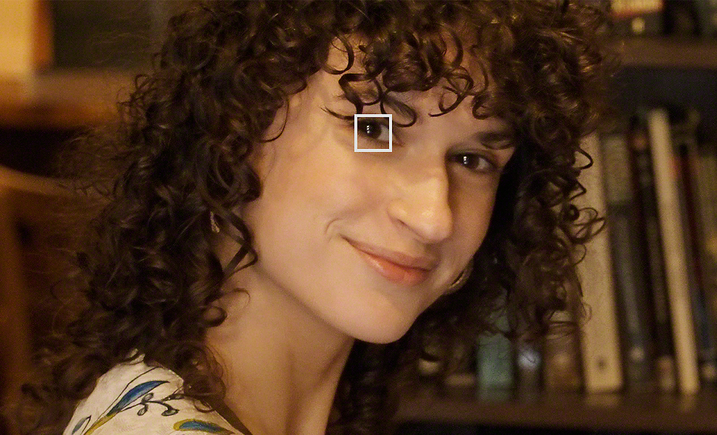 Nainen katselee kohti kameraa kirjastossa – pieni valkoinen neliö hänen toisen silmänsä päällä kuvaa videokuvauksen Eye AF -toimintoa