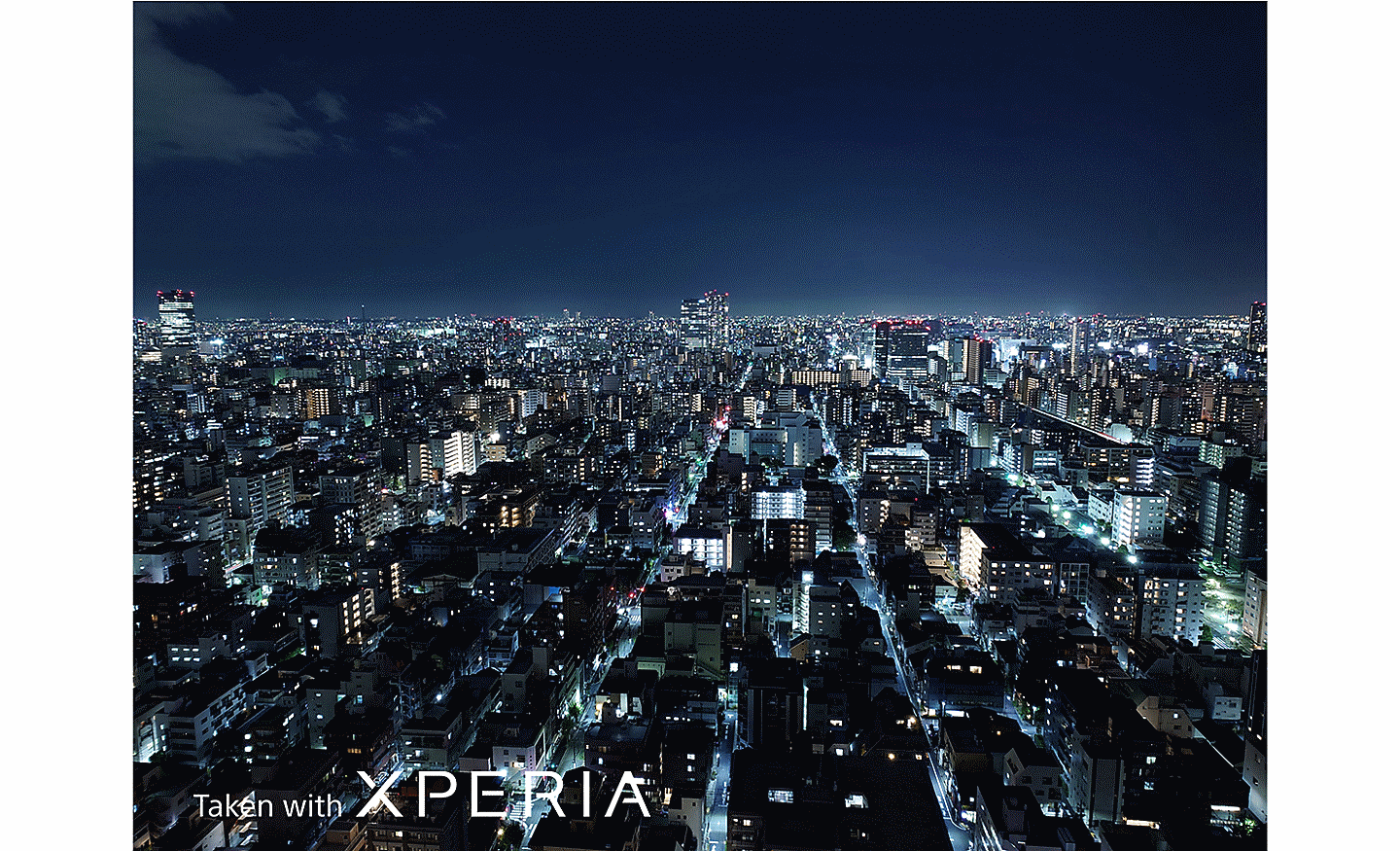Rozľahlá nočná panoráma mesta zachytená z výšky. Na fotografii sa nachádza text Taken with XPERIA.