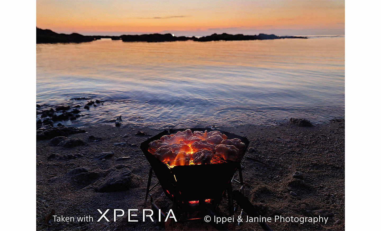 Žhnoucí ohniště na pláži s výhledem na vodu při západu slunce a text „Pořízeno smartphonem XPERIA © Ippei & Janine Photography“