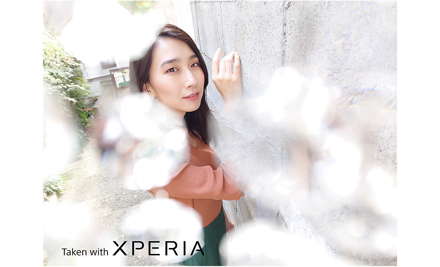 Portrait of a woman in an external setting with blurred objects in the foreground. Text reads "Taken with XPERIA".