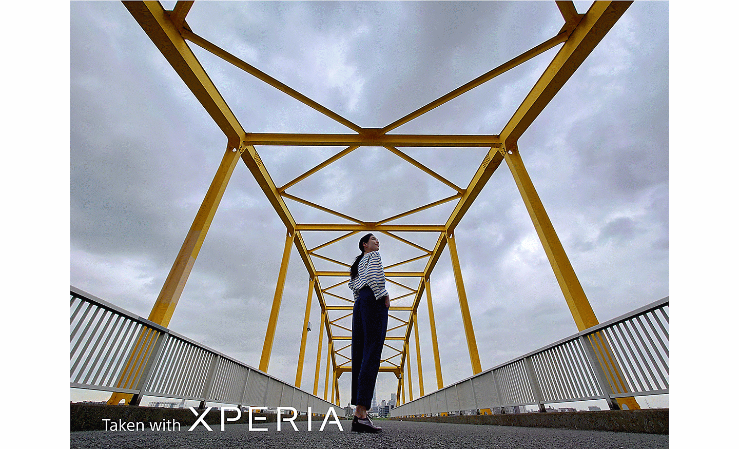 Úchvatná fotografia ženy pózujúcej na kovovom moste. Na fotografii sa nachádza text Taken with XPERIA.