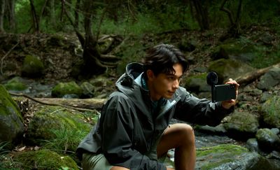 Photo of a person in a grey waterproof jacket crouching in a wooded area, holding a smartphone with an ECM-AX10 microphone to record.
