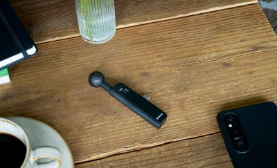 Photo of Black ECM-AX10 microphone on a wooden table beside a smartphone, coffee cup, notebook, and ribbed glass vase.