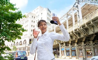 Photo of a person holding a drink and a purple smartphone with an attached ECM-AX10 microphone in front of city buildings.