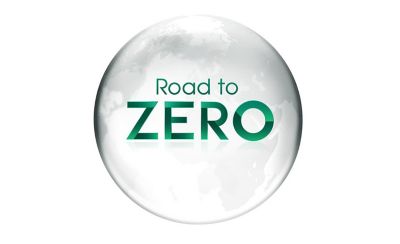 Un logotipo gráfico con el texto "Road to ZERO" centrado dentro de una esfera blanca translúcida y brillante que ilustra un mapa gris tenue de los continentes. Las palabras "Road to" están escritas en una tipografía verde delgada, justo encima de la palabra "ZERO", la cual aparece en una tipografía verde llamativa, gruesa y de estilo geométrico con un sutil sombreado degradado