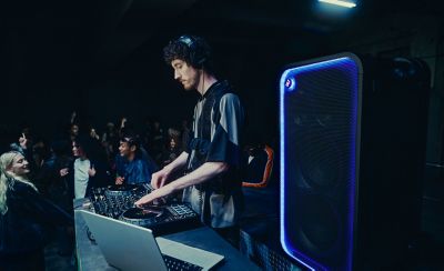 Un DJ con audífonos maneja una consola de mezcla con una laptop abierta sobre una mesa en un lugar subterráneo o discoteca con poca luz. Junto al equipo de DJ hay un gran parlante para fiestas ULT TOWER MAX de Sony negro. El contorno de la rejilla de malla está delineado por una brillante y llamativa tira de luces LED azul con un botón "ULT" brillante que se ve en la esquina superior. En el fondo se ve a una multitud bailando en la oscuridad.