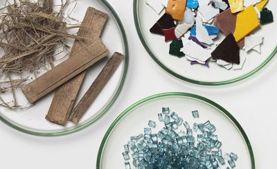 Toma cenital que muestra tres placas de Petri de cristal transparente dispuestas sobre una superficie blanca, cada una contiene diferentes materiales utilizados en la fabricación sostenible. La placa en la parte superior izquierda contiene fibras vegetales secas y piezas de madera planas y desgastadas. La placa en la parte superior derecha está llena de trozos de plástico multicolor fragmentados en amarillo, rojo, blanco y negro. La placa inferior contiene una pila de pequeños gránulos de plástico reciclado azul translúcido.
