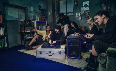Una imagen de estilo de vida que muestra a un grupo de jóvenes pasando el rato en un sótano o habitación decorados de forma informal y con poca luz. En el centro, un DJ con audífonos mezcla música en una consola colocada sobre un estuche de transporte metálico de color plateado. Justo al lado de la consola hay un parlante para fiestas ULT TOWER 5 de Sony negro. El contorno de la rejilla rectangular frontal está delineado por un brillante anillo de luz LED azul y morada. Los amigos están sentados en el piso y en los sofás, charlando y disfrutando de la música.