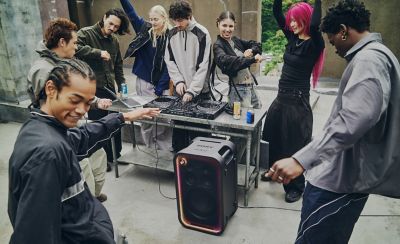 Fotografía de un grupo de personas que bailan en una azotea alrededor de un DJ. En primer plano, un parlante ULT TOWER 7 de Sony negro con un marco LED multicolor iluminado está en el piso. Al fondo, una persona con el pelo corto y rizado maneja un controlador de DJ sobre una mesa metálica.