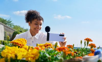 Phot of a person with ponytail holding a purple smartphone behind flowers, with an attached ECM-AX10 microphone and fluffy windscreen.