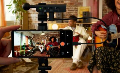 Photo of a hand-held smartphone on a tripod recording three musicians against a brick wall, equipped with a top-mounted ECM-AX10 microphone.