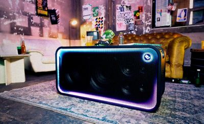 El parlante para fiestas ULT TOWER MAX de Sony está ubicado en posición horizontal sobre una alfombra estampada retro en el centro de una habitación de estilo ecléctico. La rejilla rectangular frontal está enmarcada por una brillante tira de luces LED que pasa del cian brillante a la izquierda al morado intenso y rosa a la derecha, con el botón circular "ULT" iluminado en la esquina superior derecha. En el fondo hay un sofá amarillo con botones, carteles retro pegados en una pared vieja, un sillón blanco y snacks sobre una mesa baja detrás del parlante.