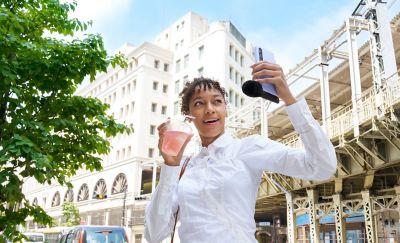 Photo of a person holding a pink drink and pointing a purple smartphone with an attached ECM-AX10 microphone on a city street.