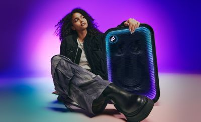 Una persona joven con el cabello rizado sentada en el piso y apoyando el brazo sobre un gran parlante para fiestas ULT TOWER 5 de Sony negro. Lleva una chaqueta negra, pantalones cargo holgados y botas negras de suela gruesa. El parlante está en posición vertical con la rejilla rectangular de malla enmarcada por una brillante tira de luces LED que pasa de cian a morado. En la esquina superior, el botón circular "ULT" está iluminado. El fondo es un estudio con una intensa iluminación degradada de color morado y azul.
