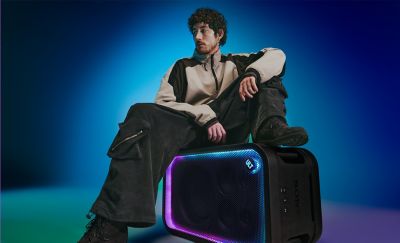 Una persona joven con el pelo rizado sentada sobre un parlante de fiesta ULT TOWER 7 de Sony negro colocado en horizontal en un estudio. Lleva una chaqueta beige y negra con cremallera, pantalones cargo negros holgados y botas oscuras mientras mira hacia un lado. La rejilla de malla rectangular frontal del parlante está enmarcada por una franja de luces LED brillantes de color púrpura y cian, con el botón circular "ULT" iluminado en la esquina superior derecha.