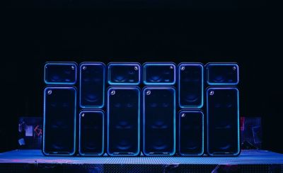 Una pared dinámica formada por los parlantes ULT POWER SOUND apilados en diferentes orientaciones verticales y horizontales. El contorno de cada unidad está delineado por brillantes anillos de luz LED azul neón sobre un fondo degradado azul y verde azulado.