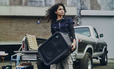 Una imagen de estilo de vida que muestra a una persona joven con el cabello rizado cargando por las asas laterales incorporadas un parlante para fiestas ULT TOWER 5 de Sony negro en posición horizontal. Está de pie al aire libre, delante de la caja de carga abierta de una camioneta clásica blanca y negra, mirando hacia un lado. La rejilla de malla frontal del parlante cuenta con un prominente botón circular "ULT" en la esquina superior. Se ve el logotipo de Sony grabado en bajo relieve en el panel de control superior.