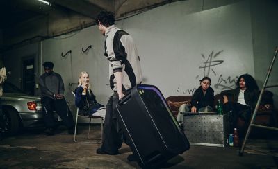 Una persona joven inclina un parlante de fiesta ULT TOWER 7 negro de Sony sobre sus ruedas y tira de él por el asa superior para desplazarlo por un garaje poco iluminado. Al fondo, un grupo de amigos conversa en un sofá y sillas cerca de un auto. Al mover el parlante, una discreta tira de luces LED púrpuras perfila la rejilla frontal.