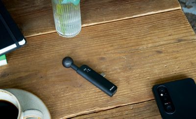 Black ECM-AX10 microphone on a wooden table beside a smartphone, coffee cup, notebook, and vase with plant stems.