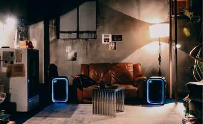 Fotografía de una habitación con un sofá de cuero marrón ubicado en el centro frente a una pared de concreto. Dos parlantes ULT TOWER 5 con iluminación LED azul en los bordes ubicados a cada lado del sofá. Delante hay una mesa de centro con listones de metal sobre una alfombra, con una lámpara de pie iluminada a la derecha.
