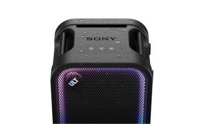 Una imagen de producto en estudio en primer plano que destaca la sección superior de un parlante para fiestas ULT TOWER 5 de Sony en negro mate cubierto de gotas de agua. La superficie de control superior muestra el logotipo "SONY" grabado en bajo relieve, las asas de transporte incorporadas y tres botones circulares al ras de la superficie, con una pequeña luz LED azul encendida arriba del botón Bluetooth. Debajo de la superficie superior húmeda, la rejilla de malla frontal cuenta con un prominente botón circular "ULT" que brilla con un degradado multicolor y está enmarcado por un anillo de luz LED activo de color morado y azul.