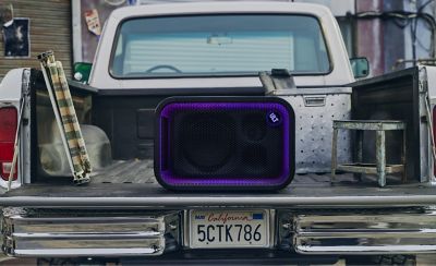 Una imagen de estilo de vida desde detrás de una camioneta retro que muestra un parlante para fiestas ULT TOWER 5 de Sony negro apoyado en posición horizontal sobre la puerta trasera abierta. La rejilla rectangular frontal del parlante está iluminada por un anillo de luz LED morada. El botón circular "ULT" brilla en la esquina superior derecha. La caja de carga de la camioneta contiene equipos para actividades al aire libre, como una silla de jardín plegada y una caja de almacenamiento de metal. Se ve una matrícula de California en el parachoques cromado en la parte inferior.