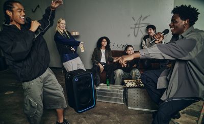 Un grupo de jóvenes se reúne en una sala decorada de forma informal, se ríen y cantan karaoke juntos. En el centro, hay un parlante de fiesta ULT TOWER 7 de Sony negro en posición vertical. El contorno de la rejilla rectangular frontal está delineado por una brillante tira de luces LED azules. Dos personas están paradas a cada lado en primer plano con micrófonos en la mano, mientras sus amigos disfrutan de la actuación sentados en un sofá al fondo.