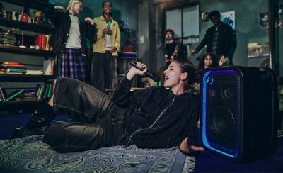 Una imagen de estilo de vida que capta a una persona joven tumbada sobre una alfombra estampada, con un micrófono en la mano y cantando con entusiasmo durante una reunión en casa. Justo a su lado hay un parlante para fiestas ULT TOWER 5 de Sony negro. El contorno de la rejilla rectangular frontal está delineado por una brillante tira de luces LED azul y morada. En el fondo poco iluminado, hay un grupo de amigos de pie sonriendo mientras ven su actuación.