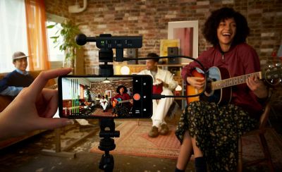 Photo of a hand-held smartphone on a tripod recording three musicians against a brick wall, equipped with a top-mounted ECM-AX10 microphone.