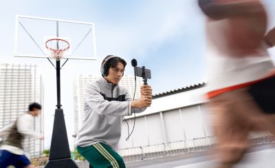 Photo of a person in grey fleece holding a smartphone with an ECM-AX10 microphone on an outdoor basketball court.