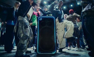El parlante ULT TOWER MAX, con un borde de LED azul brillante, se encuentra en el piso mientras la gente baila a su alrededor en una sala atmosférica con poca luz.