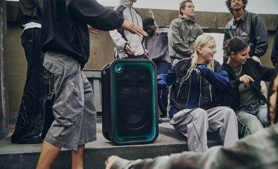 Una toma de la sección central captura a un grupo de jóvenes que pasan el rato en una superficie de cemento junto a un parlante de fiesta ULT TOWER 7 de Sony negro. El parlante está en posición vertical, con la rejilla de malla frontal enmarcada por una tira de luces LED que brilla con un brillante degradado cian y verde y un botón circular "ULT" iluminado en la esquina superior izquierda. Las personas están vestidas con ropa casual y holgada de calle, incluidos pantalones cortos holgados y chaquetas oscuras mientras bailan o se mueven alrededor de la unidad.