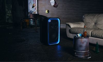 Un parlante de fiesta ULT TOWER 7 de Sony negro colocado en posición vertical sobre un fondo gris texturizado en un estudio. La rejilla rectangular frontal del parlante está enmarcada por una tira de luces LED azul brillante, con el botón circular "ULT" iluminado en la esquina superior derecha. El telón de fondo presenta una superficie azul metálica altamente reflectante con reflejos de luces verticales.