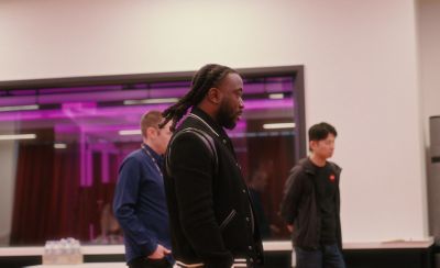 Tres personas de pie en un estudio con iluminación de tono rosa. El hombre que está en el centro, que lleva una camiseta con un estampado, hace un gesto con la mano mientras habla con el hombre a su izquierda.