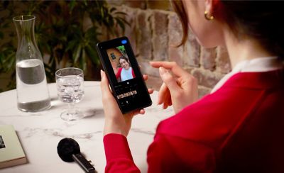 Photo of a person in a red top at a marble table editing video on a smartphone beside an ECM-AX10 microphone with windscreen resting on the table.