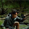 Photo of a person in a grey waterproof jacket crouching in a wooded area, holding a smartphone with an ECM-AX10 microphone to record.