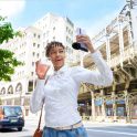 Photo of a person holding a drink and a purple smartphone with an attached ECM-AX10 microphone in front of city buildings.