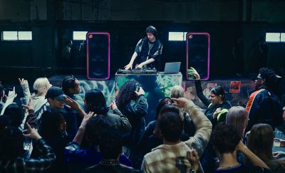 Un DJ se presenta en una cabina de mezcla ante un público que baila, rodeado por parlantes ULT TOWER MAX que brillan con un borde de iluminación LED en tonos naranja y rosa al fondo.
