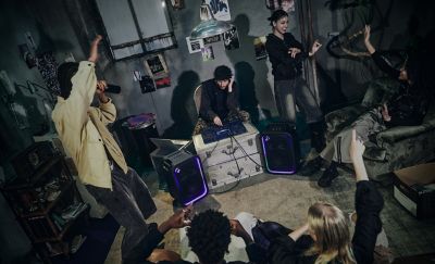 Una imagen desde arriba que muestra a un pequeño grupo de amigos disfrutando de la música en una sala de estilo industrial con poca luz. En el centro, hay un parlante para fiestas ULT TOWER 5 de Sony negro en posición vertical sobre una alfombra. La rejilla frontal está iluminada por un anillo de luz LED morada y azul. A la izquierda, un hombre maneja una consola de mezcla de DJ azul colocada sobre baúles de metal apilados. Otra mujer baila cerca, mientras una amiga se relaja en un sillón floral retro y levanta la mano para bailar con las personas sentadas en el piso que se encuentran en primer plano.