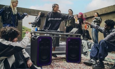 Un grupo de jóvenes se reúne en un patio de cemento al aire libre en la azotea para una fiesta con DJ. Dos parlantes de fiesta ULT TOWER 7 de Sony negros colocados verticalmente en primer plano sobre una alfombra estampada, con las rejillas frontales iluminadas por un borde LED púrpura. En el centro, varias personas bailan y socializan alrededor de una mesa de metal que sostiene una laptop y un equipo de audio mientras tintinean latas y botellas de bebidas.