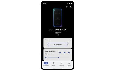 Captura de pantalla de un teléfono celular que muestra la interfaz de la aplicación complementaria Sony Sound Connect sobre un fondo oscuro. En la parte superior, se muestra una imagen del parlante ULT TOWER MAX en posición vertical. Debajo hay un texto blanco en negrita que dice "ULT TOWER MAX", seguido de un indicador de batería en "100 %" y el códec de audio "AAC". Debajo hay dos tarjetas de control de color gris claro: la tarjeta superior detalla la "función" actual como conexión "Bluetooth" y la tarjeta inferior muestra un panel de control de reproducción con los botones para retroceder, reproducir y avanzar, además de un control deslizante horizontal para ajustar el volumen. Hay una barra de navegación blanca en la parte inferior de la pantalla con íconos con las etiquetas "Mi dispositivo", "Escena", "Descubrir" y "Menú".