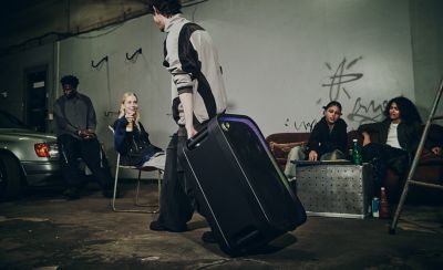 Una persona joven tira de un parlante de fiesta ULT TOWER 7 negro de Sony por el asa superior y lo inclina sobre las ruedas de transporte integradas para desplazarlo por un espacio interior poco iluminado. Al fondo, un pequeño grupo de amigos se relaja y conversa en un sofá y unas sillas vintage cerca de un auto antiguo. La rejilla rectangular frontal del parlante brilla sutilmente a lo largo de los bordes con una luz púrpura.