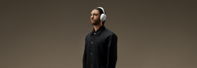 A man in a black coat stands against a brown gradient background, wearing white 1000X The Collexion headphones.