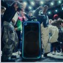 El parlante ULT TOWER MAX, con un borde de LED azul brillante, se encuentra en el piso mientras la gente baila a su alrededor en una sala atmosférica con poca luz.