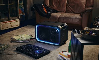 Un parlante para fiestas ULT TOWER 5 de Sony negro colocado en posición horizontal sobre una gran alfombra gris en una habitación decorada de forma informal. La rejilla rectangular de malla frontal está enmarcada por una brillante tira de luces LED azules y moradas. En el lado derecho, el botón circular "ULT" está iluminado. En primer plano, hay un controlador de mezcla de DJ negro en el piso junto a discos de vinilo apilados. El fondo está compuesto por un sofá de cuero marrón desgastado, libros y una mesita.