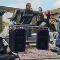 Fotografía de un DJ al aire libre sobre una alfombra estampada. Una persona de pie detrás de una mesa con estructura metálica controla el equipo, flanqueada por dos parlantes ULT TOWER 7 con iluminación LED púrpura. Otras personas se reúnen en el fondo.