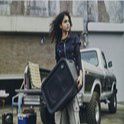 Una imagen de estilo de vida que muestra a una persona joven con el cabello rizado cargando por las asas laterales incorporadas un parlante para fiestas ULT TOWER 5 de Sony negro en posición horizontal. Está de pie al aire libre, delante de la caja de carga abierta de una camioneta clásica blanca y negra, mirando hacia un lado. La rejilla de malla frontal del parlante cuenta con un prominente botón circular "ULT" en la esquina superior. Se ve el logotipo de Sony grabado en bajo relieve en el panel de control superior.