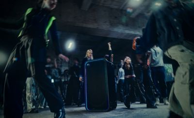 Fotografía de un parlante ULT TOWER MAX con iluminación LED azul en los bordes ubicado en el centro de una sala de concreto. Un grupo de personas baila alrededor del parlante. Hay figuras desenfocadas en primer plano y una persona con cabello rubio y largo al fondo.