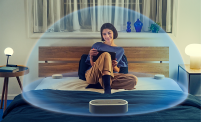 Image of a person sitting on a bed surrounded by the HT-AX7 and a transparent bubble encapsulating them with spatial sound